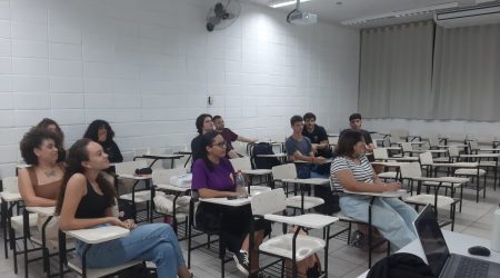 A imagem mostra uma sala de aula com onze jovens, homens e mulheres, sentadas em carteiras distribuídas pela sala. Eles estão atentos olhando para frente. Há paredes brancas e uma cortina cinza no fundo. No teto, há luminárias acesas e um projetor.