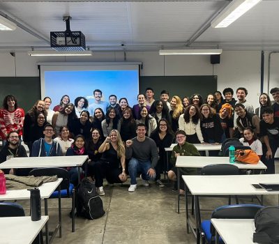 Uma sala de aula com mesas vazias e cadeiras à frente e um grupo de aproximadamente 50 pessoas posadas para foto. As duas pessoas do centro apresentam o braço erguido em comemoração.