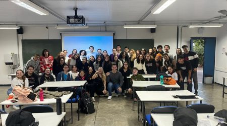 Uma sala de aula com mesas vazias e cadeiras à frente e um grupo de aproximadamente 50 pessoas posadas para foto. As duas pessoas do centro apresentam o braço erguido em comemoração.