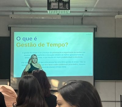 Uma mulher loira de camiseta preta fazendo uma apresentação de texto sobre gestão de tempo em uma tela de projeção. Há pessoas à frente assistindo a apresentação.