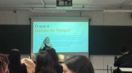 Uma mulher loira de camiseta preta fazendo uma apresentação de texto sobre gestão de tempo em uma tela de projeção. Há pessoas à frente assistindo a apresentação.