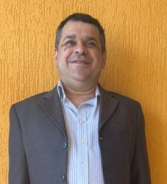 Foto de Alessandro, um homem branco de cerca de 40 anos, com deficiência visual, em plano frontal. Ele veste um terno cinza e uma camisa branca, com cabelos castanhos escuros e barba feita. O fundo da imagem é uma grande parede laranja.