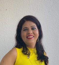 Foto vertical de Doriane Vasconcelos, mulher com cabelos escuros, lisos e cacheados nas pontas. Ela tem um rosto pouco ovalado e sorri. Doriane veste blusa amarela, com uma parede branca ao fundo.