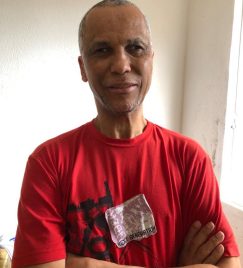 Foto de Ednilson Sacramento, homem negro de 50 anos, sem cabelos, rosto fino, sobrancelhas finas, braços cruzados, camiseta vermelha. Parede branca ao fundo.