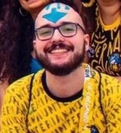 No centro da imagem, um jovem branco careca vestido com uma camiseta amarela com os escritos NAUMTERIA em preto. Ele tem barba e bigode, usa um óculos retangular com uma armação preta e está sorrindo. No topo de sua cabeça, há o desenho de uma seta azul apontada para baixo.