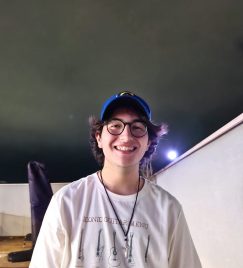 Fotografia de um homem jovem sorrindo com cabelo longo na cor preta. Ele veste uma camisa branca, um boné azul e usa um óculos com armação preta. No fundo, há uma parede branca com uma lâmpada de luz forte. O céu está nublado e escuro.