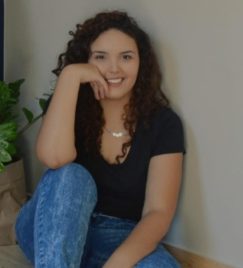 Foto de uma moça com cabelos castanhos, longos e ondulados, sorrindo para a câmera. Ela veste uma camiseta preta e um colar com pingente circular prateado. Ao fundo, destaca-se uma parede branca.