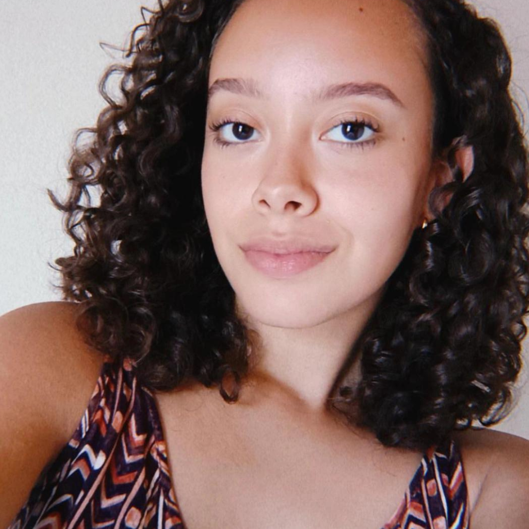 Fotografia selfie de Isabella, jovem de pele clara e sorriso cativante. Cabelo cacheado castanho escuro, blusa com estampa geométrica. Plano frontal, fundo com parede branca.