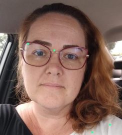 Selfie de Leila Felipini, mulher branca com cabelo tonalizado em castanho claro, olhos castanhos e sobrancelhas da mesma cor. Ela está dentro de um carro, usando uma camiseta preta e branca e óculos com armação vermelha.