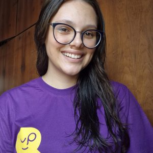 Fotografia de uma mulher jovem com cabelos e olhos castanhos. Usa óculos de grau e está sorrindo para a foto. Veste a camiseta roxa do Biblioteca Falada. O fundo da foto é uma porta de madeira.