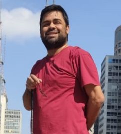 Paulo Henrique, homem branco com deficiência visual, em foto de frente. Cabelo curto e liso, barba curta. Sorri, mão direita para trás, mão esquerda para frente. Camisa vinho. Fundo: céu azul e prédios ao redor.