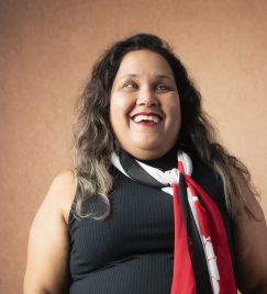 Cristiane Rufino, mulher com deficiência visual, em foto de frente. Cabelos pretos com mechas loiras caem sobre os ombros. Rosto ovalado, sobrancelhas finas. Maquiagem leve, batom vermelho. Veste blusa preta, cachecol branco e vermelho. Fundo: parede marrom.