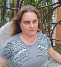 Fotografia de Juliana Peixoto. Juliana é uma mulher branca com deficiência visual com seus 40 e poucos anos de idade. Ela tem cabelos em tom de loiro escuro, rosto oval e traços finos. Juliana usa uma camiseta cinza e shorts amarelo. Ela está sentada um banco de madeira. Ao fundo, podemos ver árvores e casas.